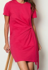 Robe fuchsia à manches courtes avec un détail noué sur le côté gauche. Fabriquée en tissu doux, mi-genou, avec une coupe décontractée et une texture lisse.