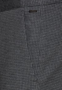 Tessuto a motivo houndstooth grigio e nero con una piccola etichetta rettangolare nera cucita vicino al centro superiore.
