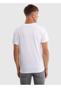 Fiatal férfi rövid barna hajjal, egyszínű fehér rövid ujjú pólót és szürke farmert viselve, háttal áll egy egyszerű háttér előtt.