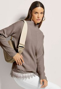 Brauner Strickpullover mit strukturiertem Design, hohem Kragen und Spitzensaum, kombiniert mit hellen Hosen und einer beigefarbenen Umhängetasche.