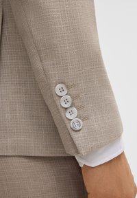 Beige strukturierte Blazerärmeln mit vier weißen Knöpfen und sichtbaren Nähdetails; ein weißes Hemdmanschette schaut unter dem Ärmel hervor.