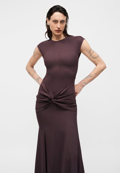 Ženska s tetovažami, oblečena v oprijeto temno vijolično obleko z vozelnim detajlom v pasu, stoji z rokami na bokih na enobarvni podlagi.