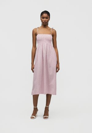 Kvinde iført en ærmeløs midi-kjole med pink og hvide striber, spaghetti stropper og flæsedetalje, kombineret med hvide højhælede sandaler.