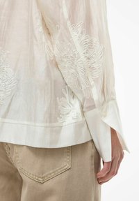 Blouse légère et transparente en ivoire pâle, ornée d'une broderie florale blanche délicate, manches longues avec détail de poignet boutonné. Associée à un pantalon beige.