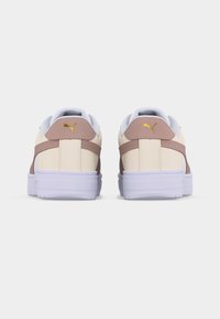 Ζευγάρι κρεμ και ανοιχτό καφέ αθλητικά παπούτσια Puma, εμφανιζόμενα από πίσω, με χρυσό λογότυπο Puma στο τακούνι και άσπρες μαλακές σόλες.