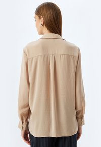 Blusa beige a maniche lunghe in tessuto liscio, con colletto classico e polsini abbottonati, caratterizzata da una piega sul retro per un maggiore drappeggio.
