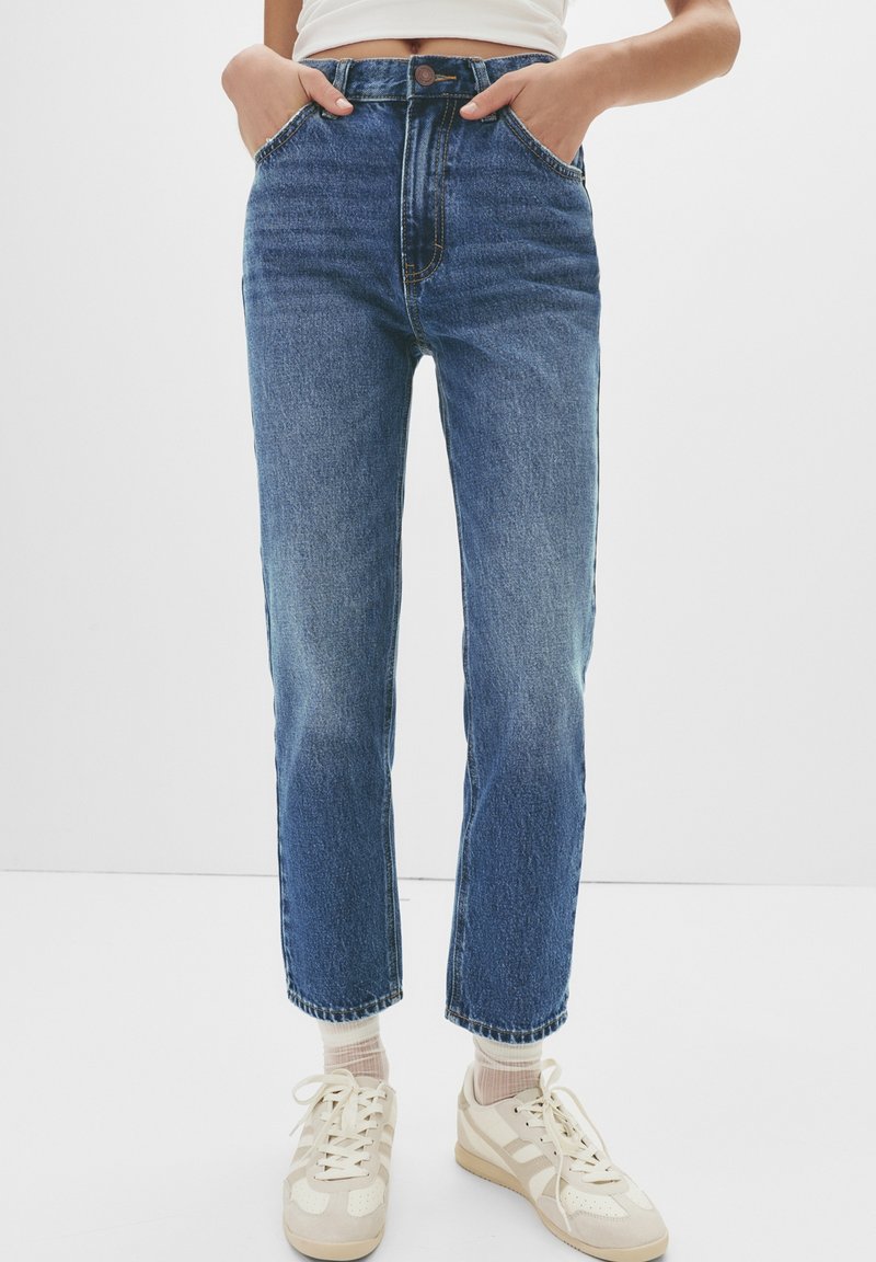 Personne portant un jean bleu taille haute, des chaussettes blanches, des baskets crème et un crop top blanc avec les mains dans les poches.