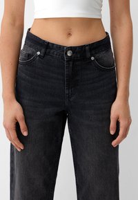 Jeans de mezclilla negra de cintura alta, con bolsillos frontales y cierre de botón. La tela tiene un acabado ligeramente texturizado, con un ligero efecto de desvanecimiento.