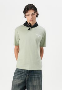 Lys grøn bomulds t-shirt med korte ærmer og et trykt "Palm Coast" logo. Sat sammen med mørkeblå jeans og et sort mønstret halstørklæde.