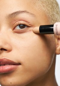 Stick correcteur crémeux dans un étui noir et rose doux appliqué sous l'œil. La peau apparaît lisse, avec un teint naturel et un éclat subtil.