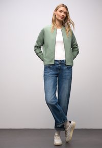 Giacca trapuntata verde chiaro con collo alla coreana, indossata sopra una camicia bianca e abbinata a jeans blu e sneakers bianche. Tessuto liscio, vestibilità casual.
