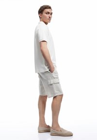 Joven de pie de lado, vistiendo una camisa polo blanca, pantalones cortos de cargo beige y zapatos de lona en una escena con fondo blanco.