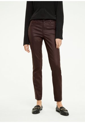 Personne portant un pantalon slim marron foncé, un haut à manches longues noir et des mocassins noirs avec une boucle dorée, debout sur un fond blanc.