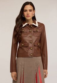Donna con lunghi capelli scuri che indossa una giacca di pelle marrone con grandi bottoni dorati e colletto bianco, abbinata a una gonna a scacchi con pieghe rosse.