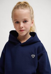 Marinblå hoodie tillverkad av mjukt tyg, med en stor huva och en liten broderad logotyp på vänstra bröstet. Minimalistisk design.