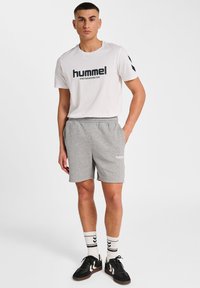 Homme debout portant un t-shirt blanc "hummel", un short gris, des chaussettes blanches à rayures noires et des chaussures de sport noires, sur un fond blanc uni.