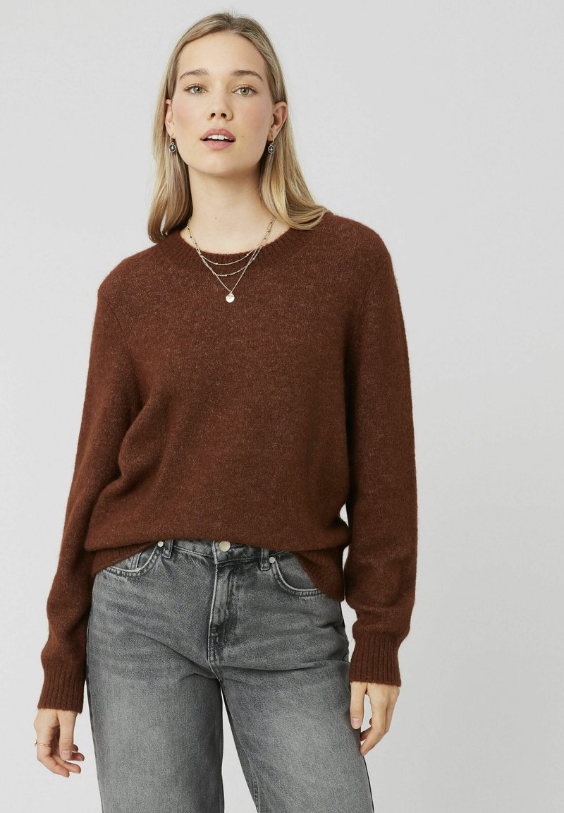Pull en tricot marron avec un ourlet et des poignets côtelés, associé à un jean gris clair. Le tissu semble doux et texturé, idéal pour un look décontracté.