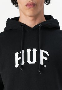 Sort hættejakke med snøre, med fed hvid "HVUF" tekst. Blødt stof, afslappet design og standard kængurulomme.