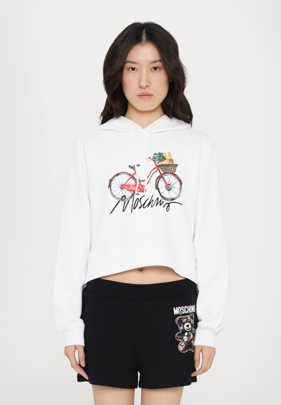 Fehér, cropped kapucnis pulcsi kerékpár grafikával és "Moschino" felirattal. Fekete rövidnadrággal párosítva, amelyen medve minta és logó részlet található.