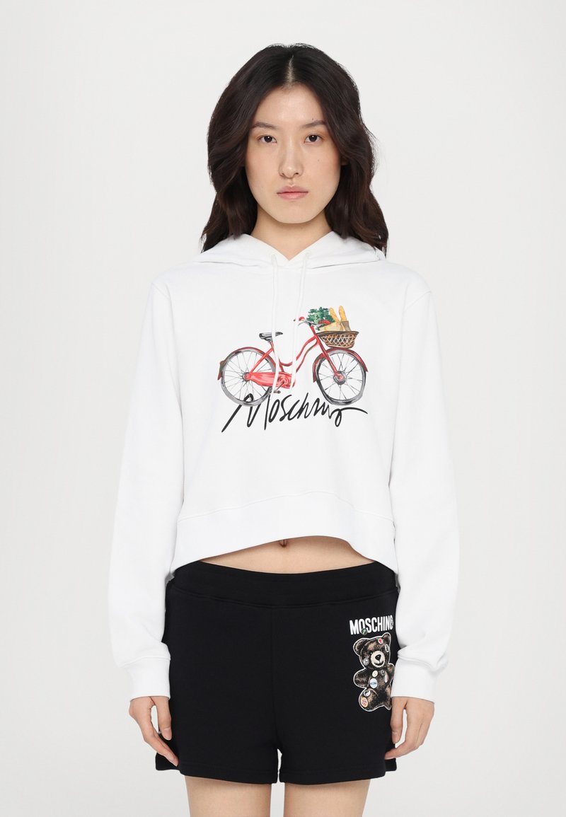 White cropped hoodie with a bicycle graphic and "Moschino" text. Paired with black shorts featuring a bear design and logo detail.