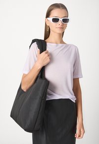 Mustat tikattu laukku, jossa on pehmustettu muotoilu, pehmeä tekstuuri ja leveä olkahihna. Mallilla on päällään vaalea violetti toppi ja valkoiset aurinkolasit.