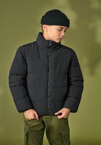 Doudoune noire avec des coutures horizontales, col montant et fermeture éclair à l'avant. Portée avec un pantalon cargo vert olive et un bonnet en tricot noir.
