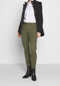 Svart quiltad jacka med huva, vit skjorta, olivgröna figursydda byxor och svarta klackade ankelstövlar. Enkel, strukturerad design och färger.