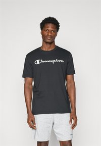 Schwarzes Baumwoll-T-Shirt mit einem weißen "Champion"-Logo auf der Brust. Kurze Ärmel, Rundhalsausschnitt, präsentiert mit grauen Shorts.
