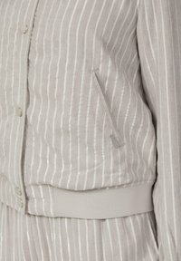 Helle graue Jacke mit vertikalen silbernen Streifen, geripptem Saum, Druckknöpfen und einer Seitentasche mit glatter Textur.