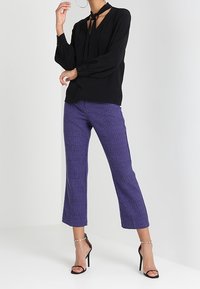 Blusa nera a maniche lunghe con cravatta al collo, abbinata a pantaloni cropped viola a righe e sandali neri con tacco.