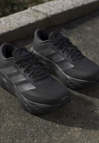 Pair of black Adidas running shoes with three dark stripes, textured soles, and laces, placed on asphalt near a stone curb.