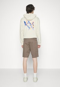 Haut à capuche crème avec un design graphique dans le dos, associé à un short marron. Modèle portant des chaussettes blanches et des baskets claires.