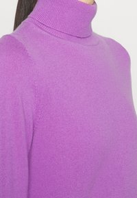 Lilla tætsiddende sweater med høj hals lavet af blødt stof, med et figursyet design og ribbede kanter ved halsen og ærmerne.