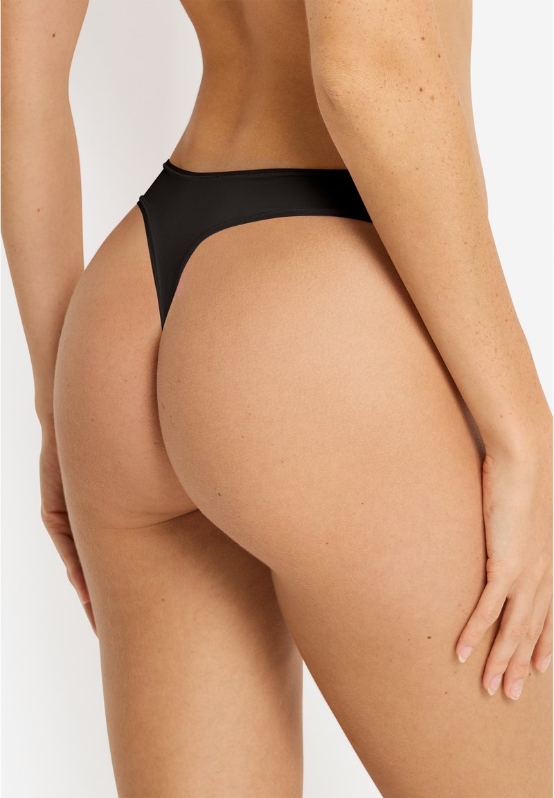 Γυναίκα με μαύρο στρινγκ, εμφανίζεται από πίσω σε ύψος μέσης και γοφών πάνω σε απλό φόντο.