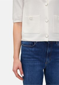 Zbliżenie na rękę osoby na kieszeni niebieskich dżinsów, noszącej białą bluzkę z guzikami i kieszeniami naszytymi.