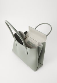 Zelena usnjena torba za čez ramo s gladko teksturo, dvema ročajema in odprtim vrhnjim delom, ki razkriva bež notranjost in žep z zadrgo.