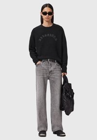 Man wearing black sunglasses, black AllSaints sweater, grey faded wide-leg jeans, black belt, black shoes, holding a black quilted bag.