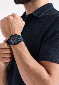 Melna analogā rokas pulksteņi ar sarkaniem akcentiem, apaļa korpusa forma, melns teksturēts ciparnieks un silikona josta. Modelis valkā tumši zilu polo krekls.