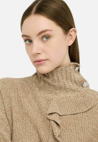 Maglione marrone a maglia con colletto alto, caratterizzato da una texture a coste e un bordo arricciato, accentuato da due decorazioni in cristallo sul colletto.