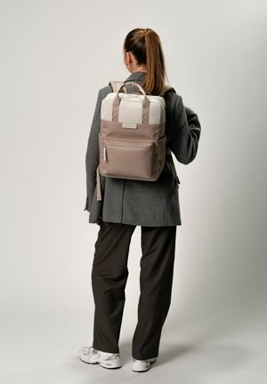 Stylischer beiger und weißer Rucksack mit gepolsterten Trägern, einem oberen Griff, einer Vordertasche und glatter Textur, getragen von einer Person in smarter Freizeitkleidung.
