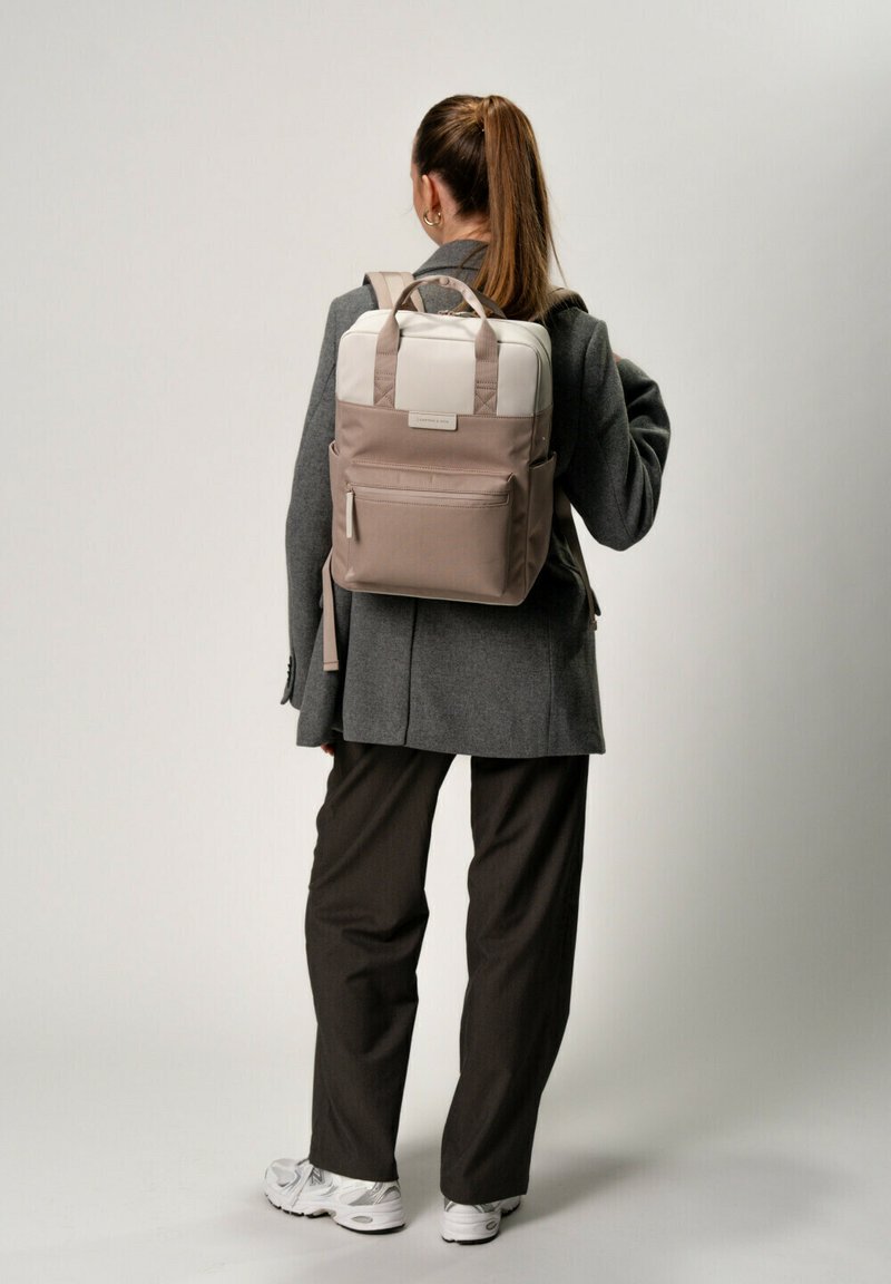 Zaino elegante beige e bianco con spallacci imbottiti, maniglia superiore, tasca frontale e texture liscia, indossato da una persona in abbigliamento smart casual.