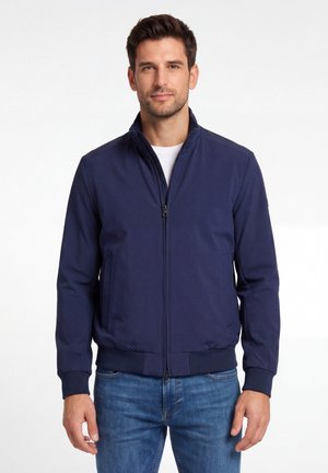 Mężczyzna w granatowej kurtce na zamek błyskawiczny, białej koszulce i niebieskich dżinsach, stojący na tle jednolitego białego tła.