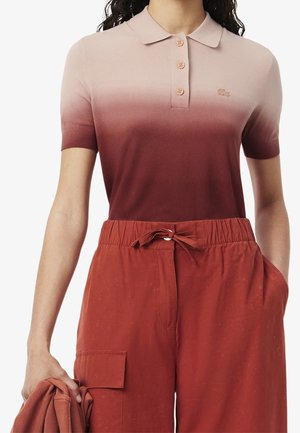 Femme portant un polo à manches courtes ombré allant du rose clair au bordeaux et un pantalon couleur rouille avec une taille à cordon.