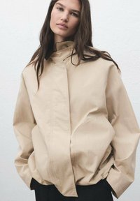 Beige oversized jakke lavet af letvægtsstof, med høj krave, skjult frontlukning og forlommer. Glat tekstur.