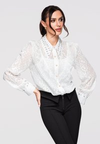 Femme posant dans un chemisier blanc transparent brodé avec col à volants et pantalon noir taille haute sur fond gris clair.