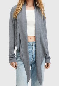 Szary prążkowany kardigan z otwartym przodem, wydłużonym dołem i pełnymi rękawami, nałożony na biały krótki top i zestawiony z jasnoniebieskimi dżinsami.