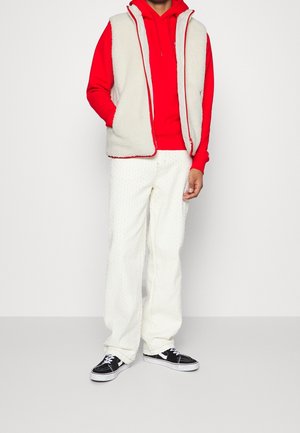 Kremowa kamizelka z polaru na czerwonej bluzie z kapturem, zestawiona z białymi teksturowanymi spodniami i czarnymi tenisówkami. Posiada zamek błyskawiczny i kontrastujące obszycia.