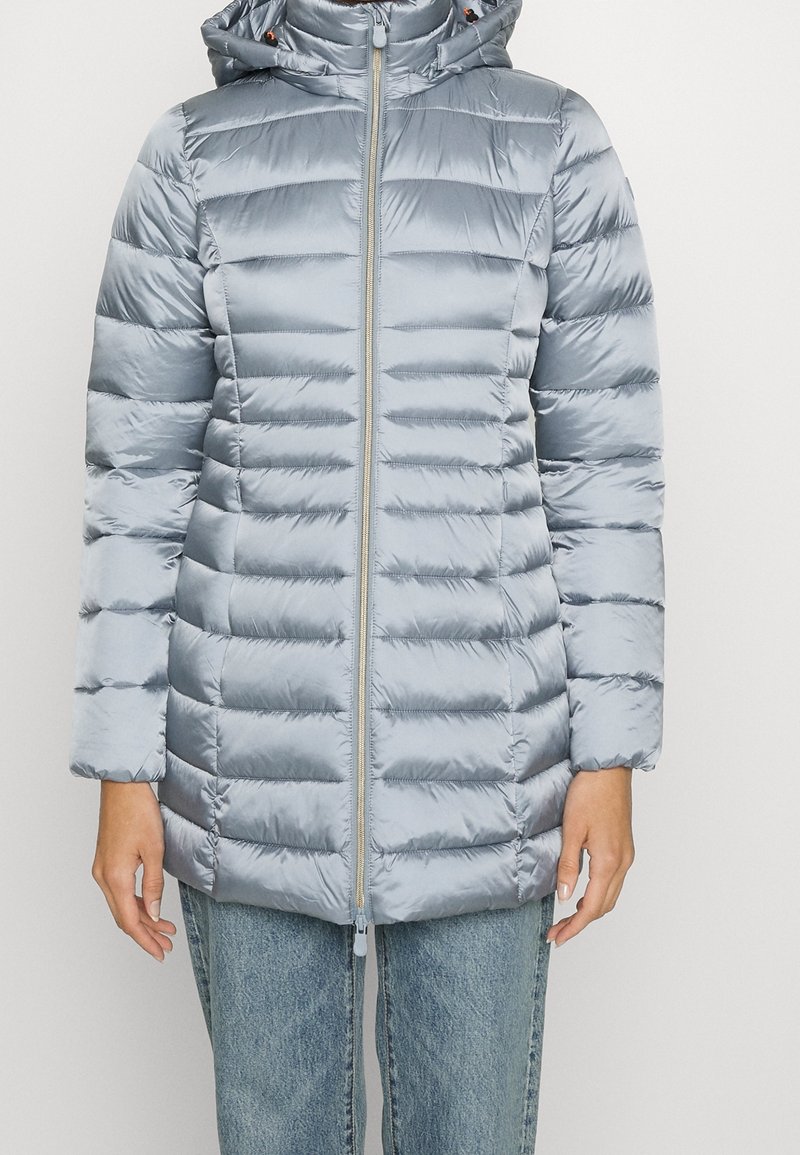Persona vistiendo una chaqueta acolchada con capucha de color azul claro, abrochada, combinada con jeans de mezclilla azul y de pie contra un fondo liso.