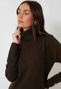 Brun ribbet turtleneck sweater med en afslappet pasform. Den strikkede tekstur har en høj krave og detaljer ved siden af, som tilføjer visuel interesse.