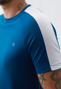 Mėlyna sportinė marškinė su trumparankoviais, pasižyminti faktūrine medžiaga, baltais peties akcentais ir maža logotipo etikete krūtinėje.